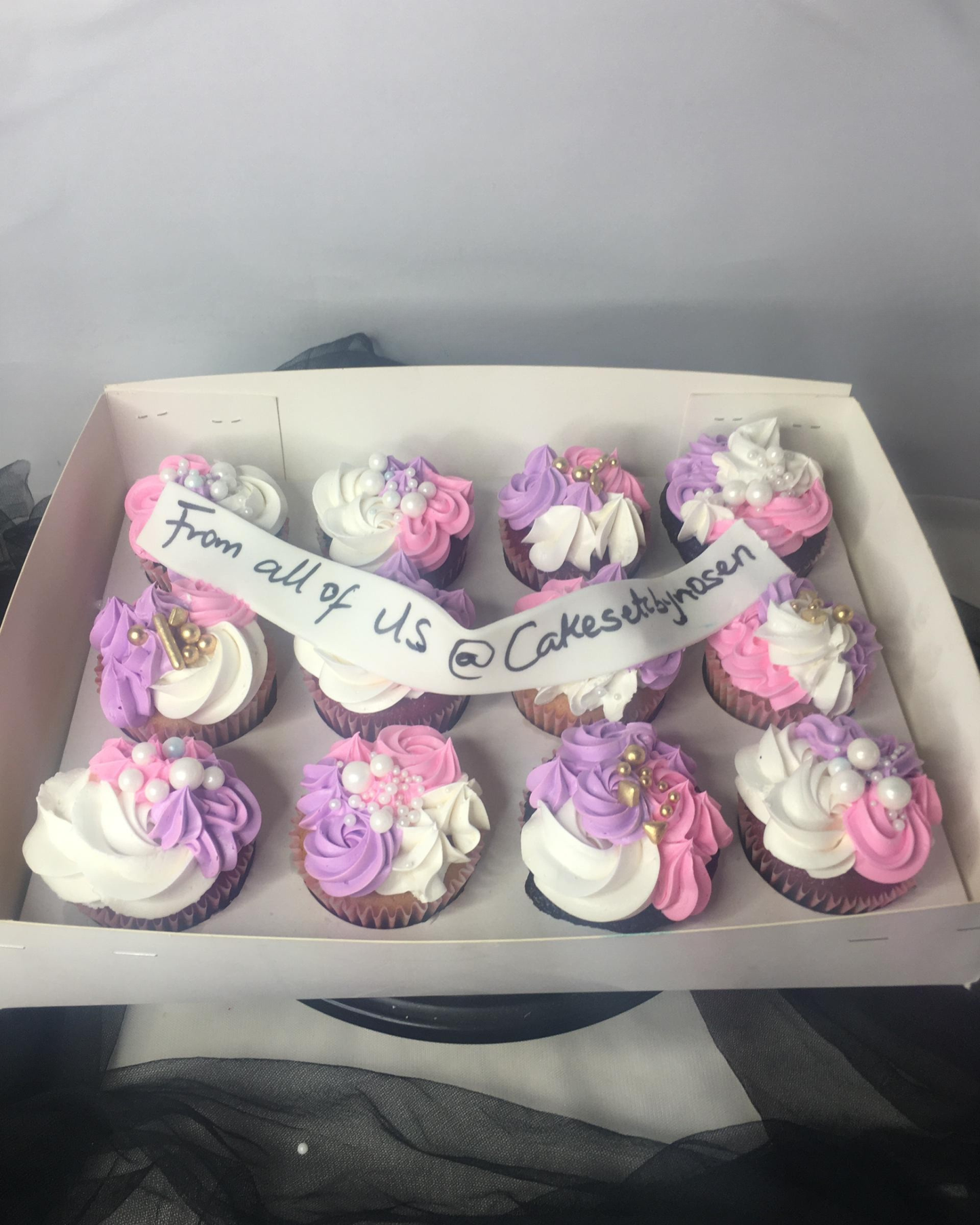 Box of cupcakes with pink, purple, and white frosting, featuring a handwritten message.