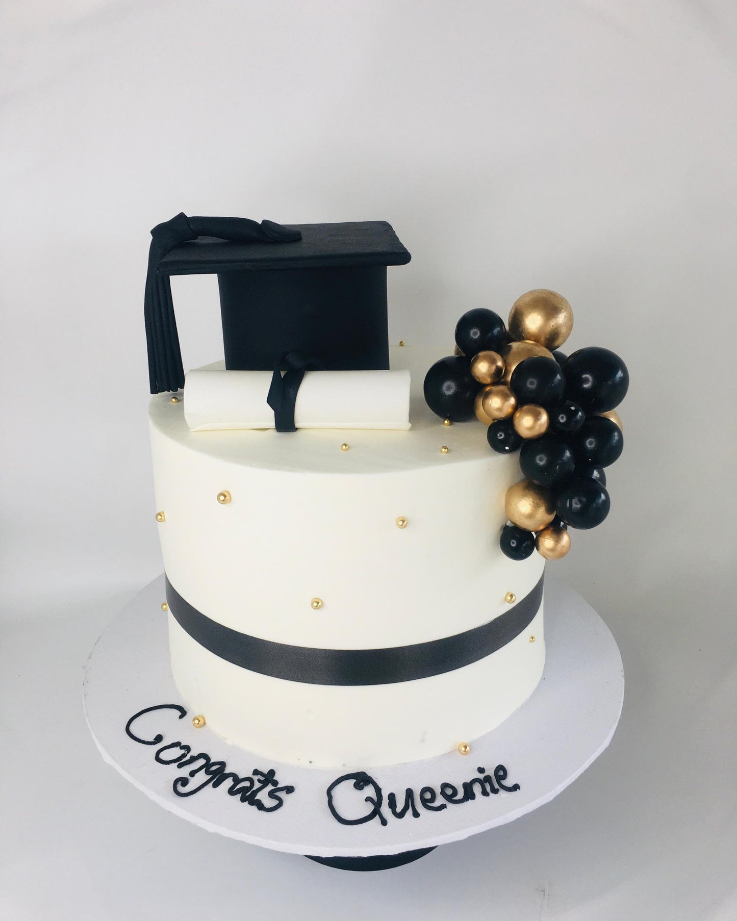 Graduation-themed cake with a black cap, gold and black balloons, and 'Congrats Queenie' text on a white background.