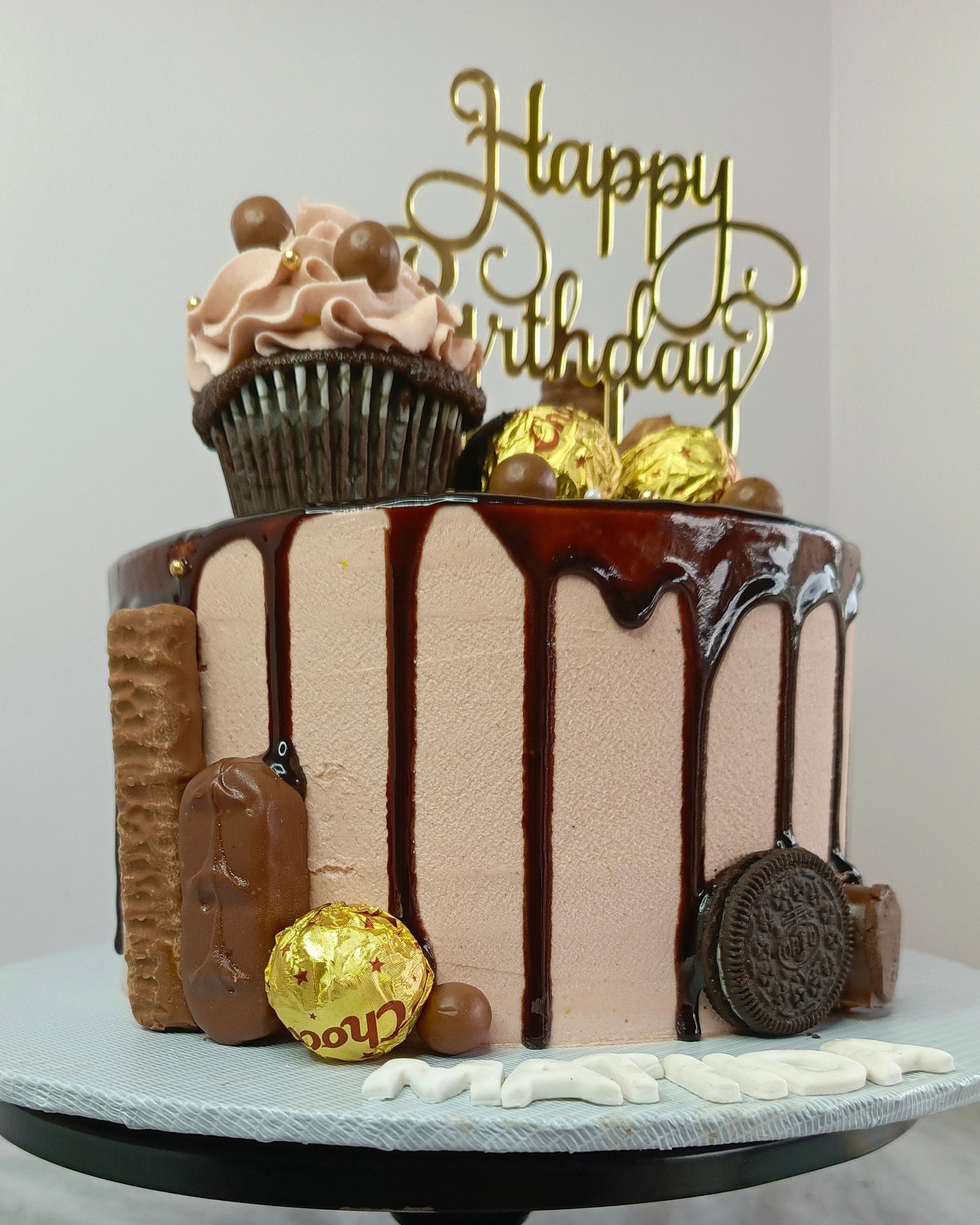 Chocolate birthday cake with decorative elements on a white stand against a light gray background.