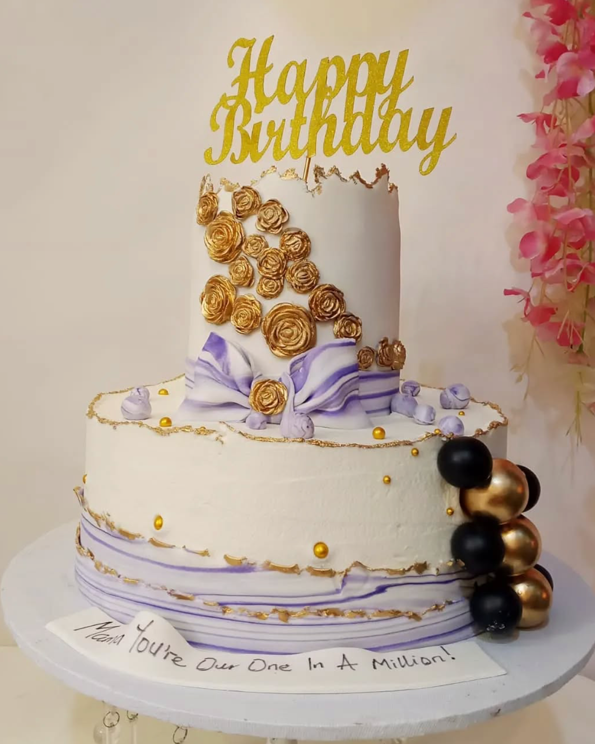 Two-tier birthday cake with gold and purple decorations, 'Happy Birthday' topper, and floral arrangement.