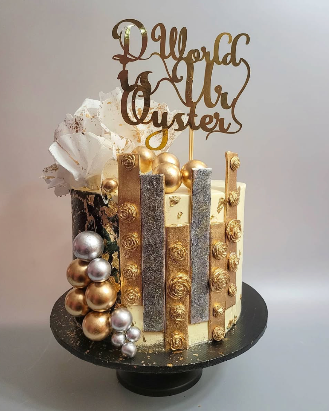 Decorative cake with gold and silver decorations and a 'The World is Our Oyster' topper on a white background.