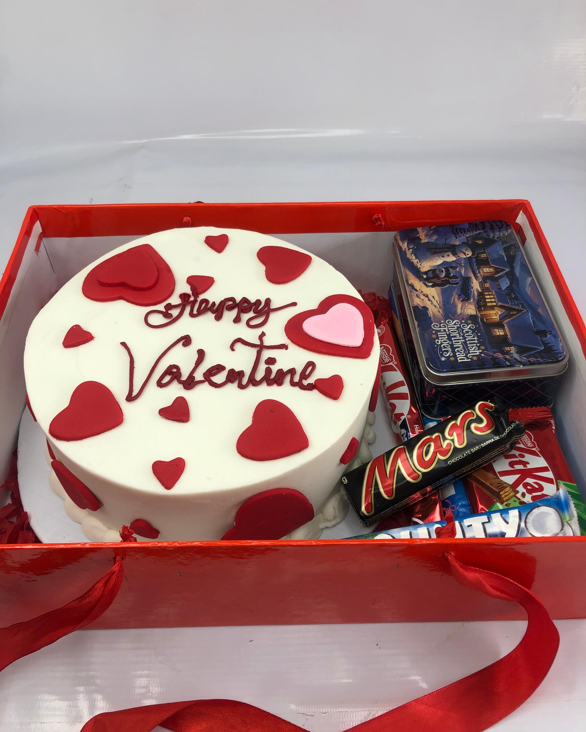 Valentine's Day cake with red hearts and 'Happy Valentine' text, surrounded by candy bars in a red box.