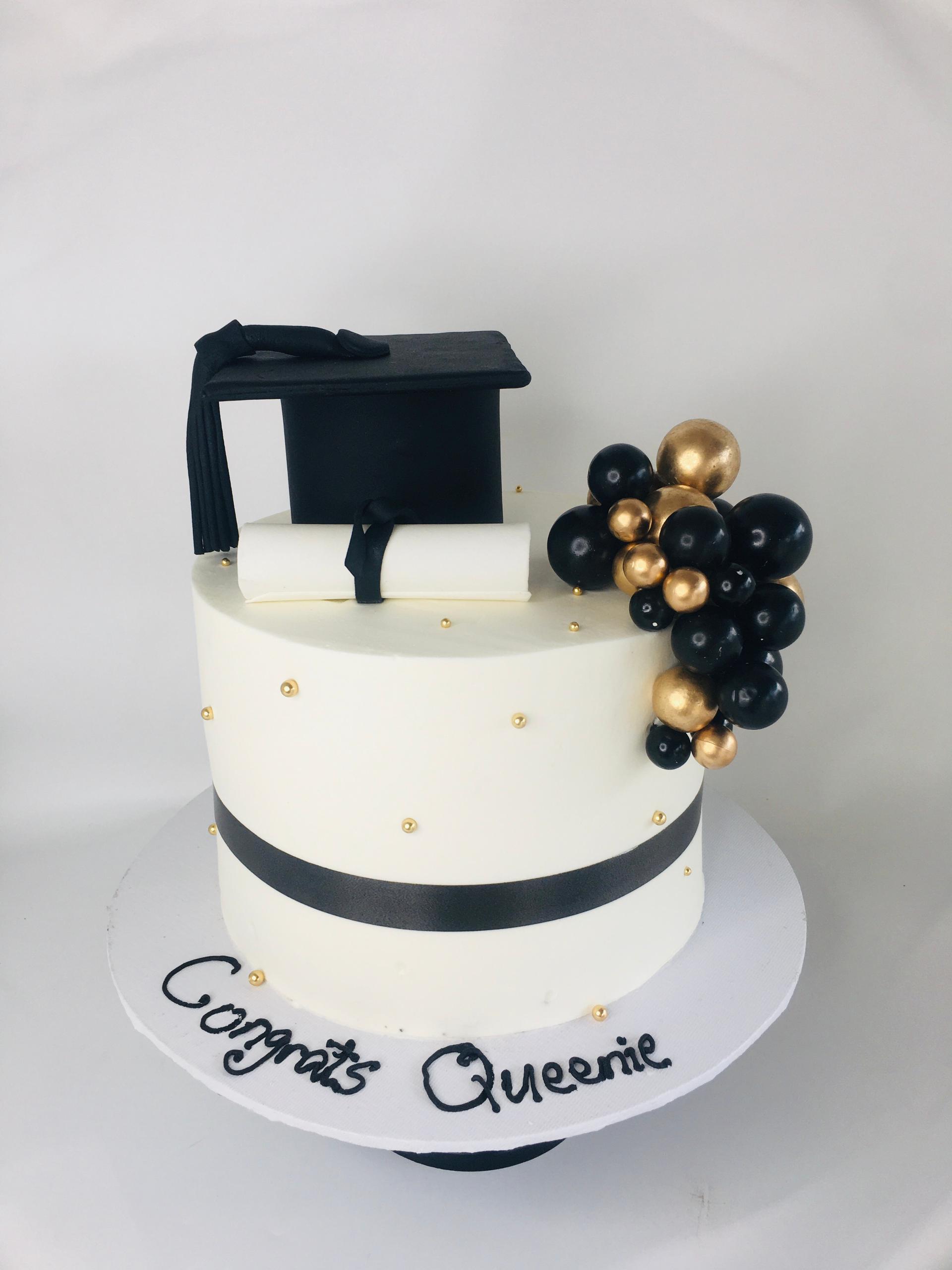 Graduation-themed cake with a black cap, gold and black balloons, and 'Congrats Queenie' text on a white background.