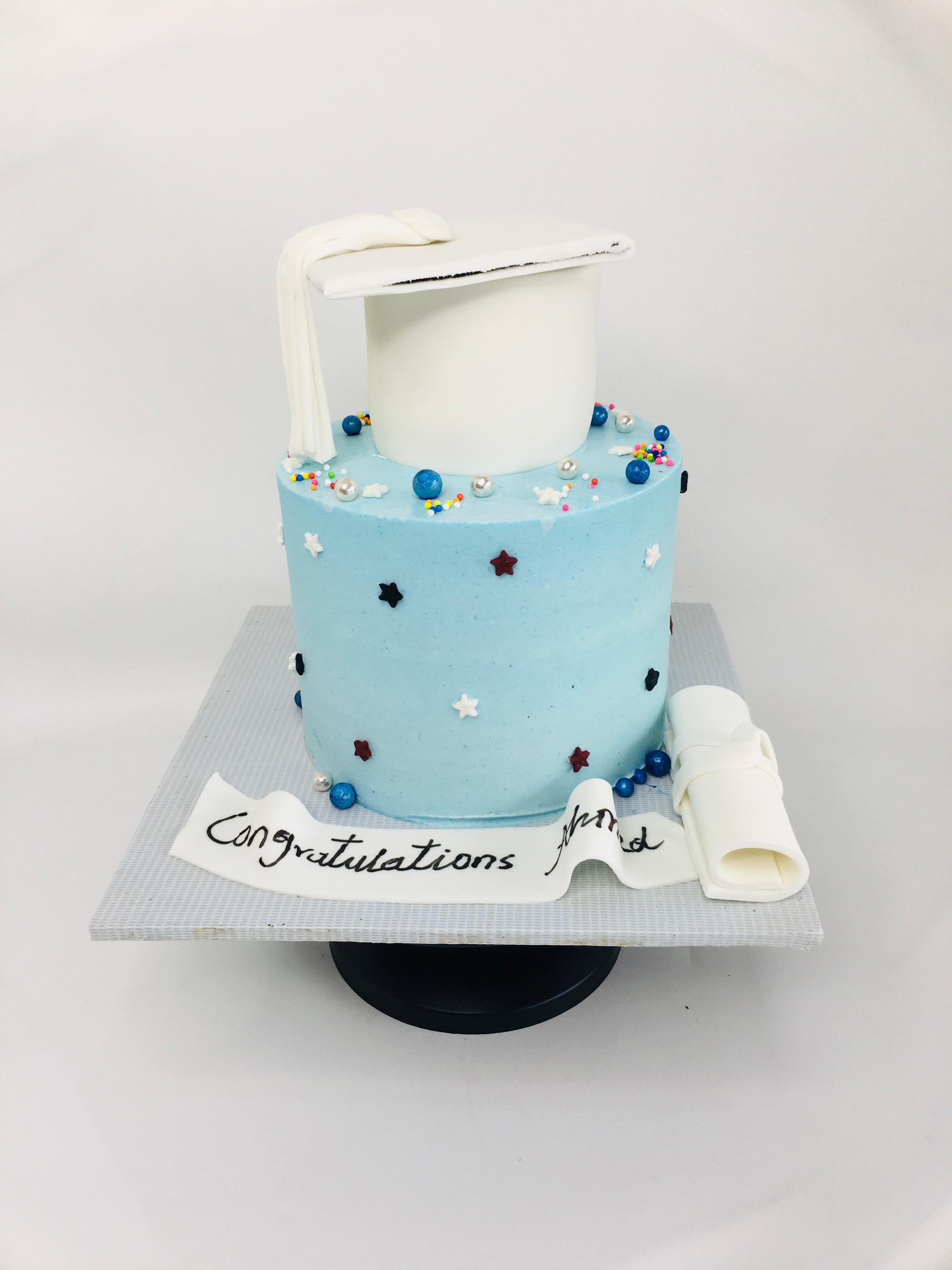 Graduation-themed cake with blue frosting, white diploma, and colorful decorations on a white background
