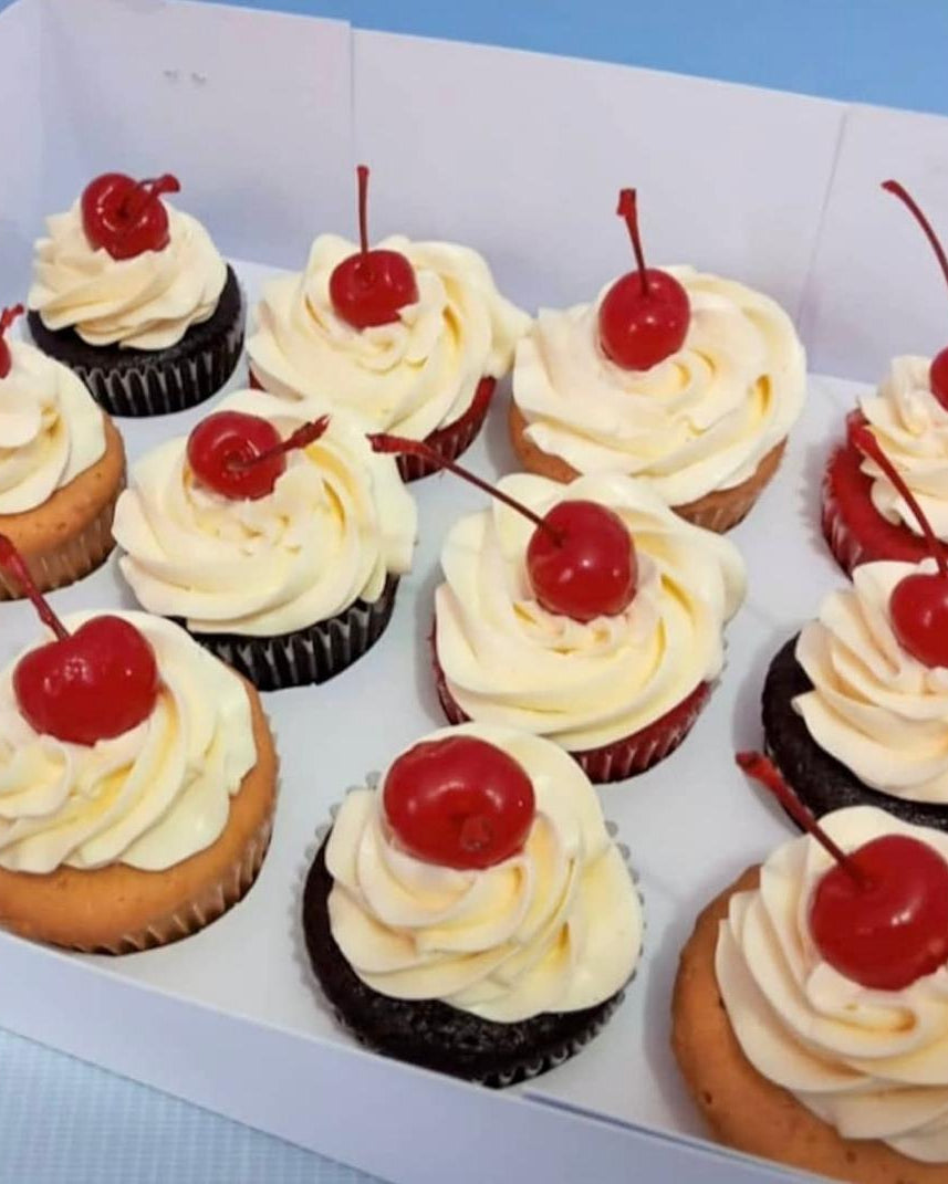 Assorted cupcakes with white frosting and red cherries in a box.